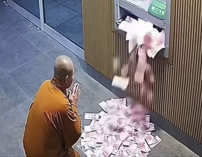 A Buddhist monk in an orange robe kneels in prayer as a large pile of paper money scatters across the floor, apparently dispensed in error from an ATM.