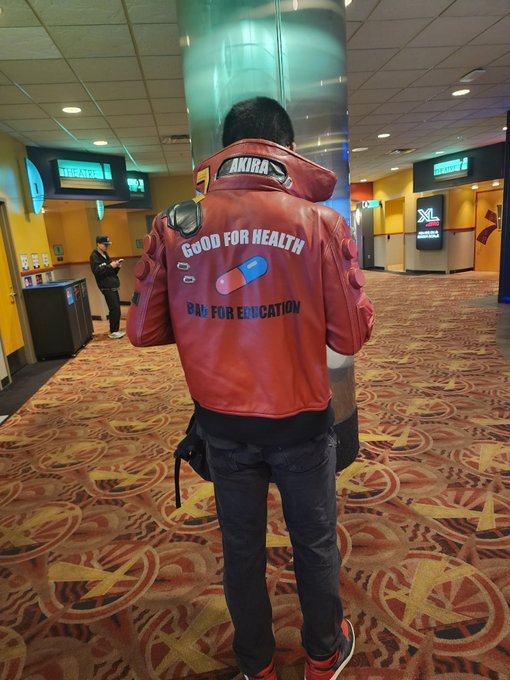 A person stands in a movie theater lobby wearing a red Akira-branded jacket with the text "GOOD FOR HEALTH" and "BAD FOR EDUCATION" alongside a pill capsule graphic on the back, with XL theater signage visible in the background.