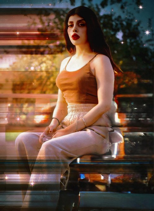 A photo of a young woman with long dark hair and red lipstick, wearing a brown tank top and beige pants, sitting outdoors with warm sunlight and a glowing sparkle filter applied to the image.