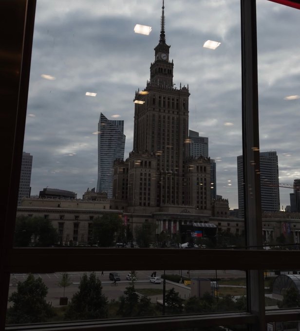 The image shows a view of the Palace of Culture and Science (Pałac Kultury i Nauki) in Warsaw, Poland, framed through a window with reflections of overhead lights visible on the glass. The surrounding modern skyline and street scene below are also visible, suggesting the photo was taken from an elevated office or workspace, matching the post's reference to tweeting from a business-focused location.