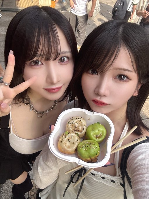 Two young women with dark hair pose together for a selfie, one making a peace sign, while holding a flower-shaped container filled with colorful buns/dumplings on a city street.