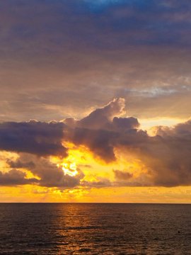 A landscape photograph of a dramatic sunset over the ocean, with bright golden sunlight piercing through dark, billowing clouds and reflecting on the water's surface.