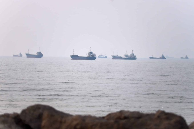 A hazy, foggy seascape showing multiple cargo ships and vessels anchored on the horizon, viewed from behind some rocks in the foreground.