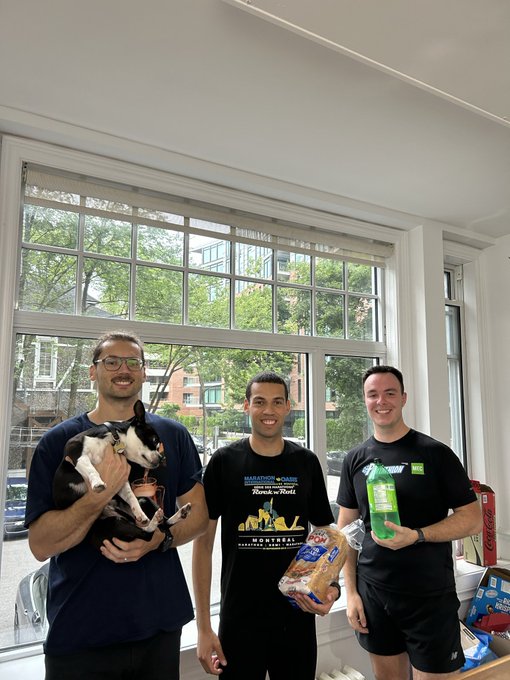 Three men pose indoors near a large window, one holding a small dog, another wearing a Montreal t-shirt and holding bread, and the third holding a green snack package.