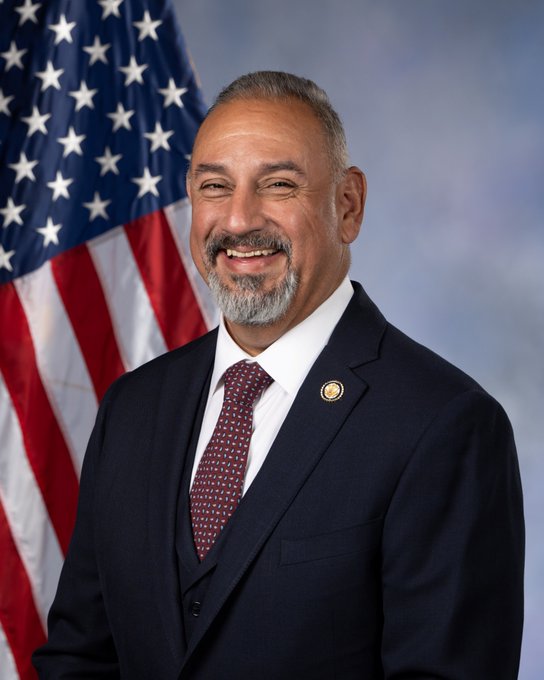 An official portrait of Rep. Gil Cisneros, smiling in a dark suit with a red patterned tie and a congressional lapel pin, posed in front of an American flag. The image accompanies commentary highlighting his timely purchase of Palantir ($PLTR) stock shortly before its 29% rise, raising questions about potential insider trading despite his position on the Armed Services Committee.