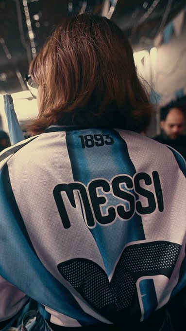 Back view photograph of a person with brown hair wearing a light blue and white striped Argentina football jersey with 'MESSI' and the number '1893' printed on the back.