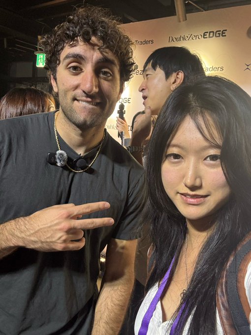 A selfie of a curly-haired man making a peace sign and a woman with long dark hair posing together at an event with a 'DoubleZero EDGE' and 'Traders' branded backdrop.
