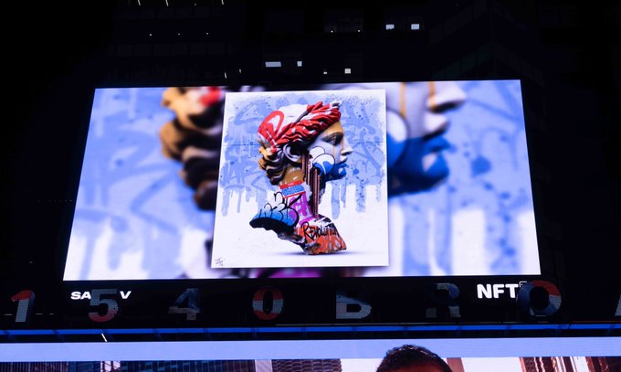 A large outdoor digital billboard displaying NFT artwork, featuring a colorful spray-can-style cartoon illustration of a lion-headed figure in red and blue.