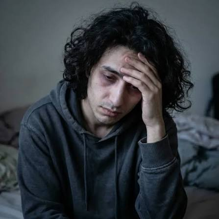 A man with disheveled curly hair and facial bruises sits on a bed, holding his head in apparent distress.