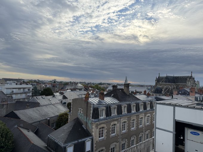 An elevated view over a city with traditional rooftops and a Gothic cathedral under a dramatic sky filled with scattered clouds.