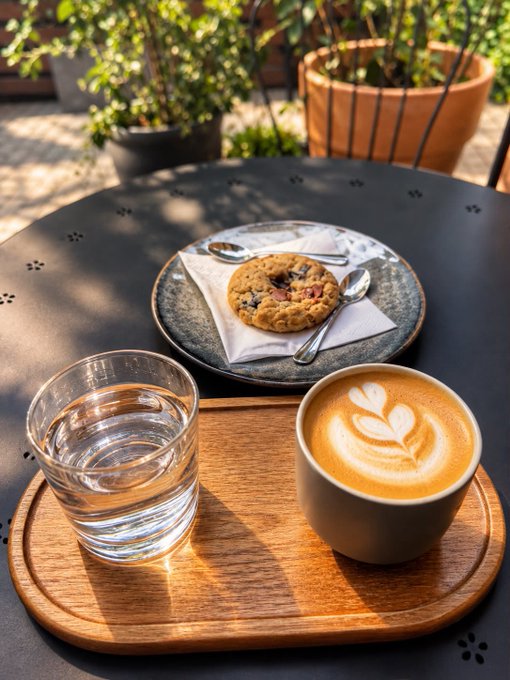 A cappuccino with leaf latte art and a glass of water are served on a wooden tray alongside a plate with a chocolate chip cookie on an outdoor cafe table.