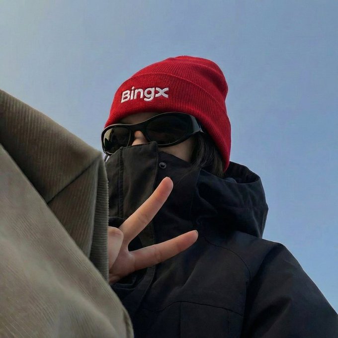 A person in a red beanie with "BingX" embroidered on it, sunglasses, and a black face-covering jacket flashes a peace sign against a clear blue sky—visually matching the caption "wearing red so the charts don't have to" by pairing a bold red accessory with the BingX brand, humorously suggesting they dressed in red to compensate for uneventful market performance.