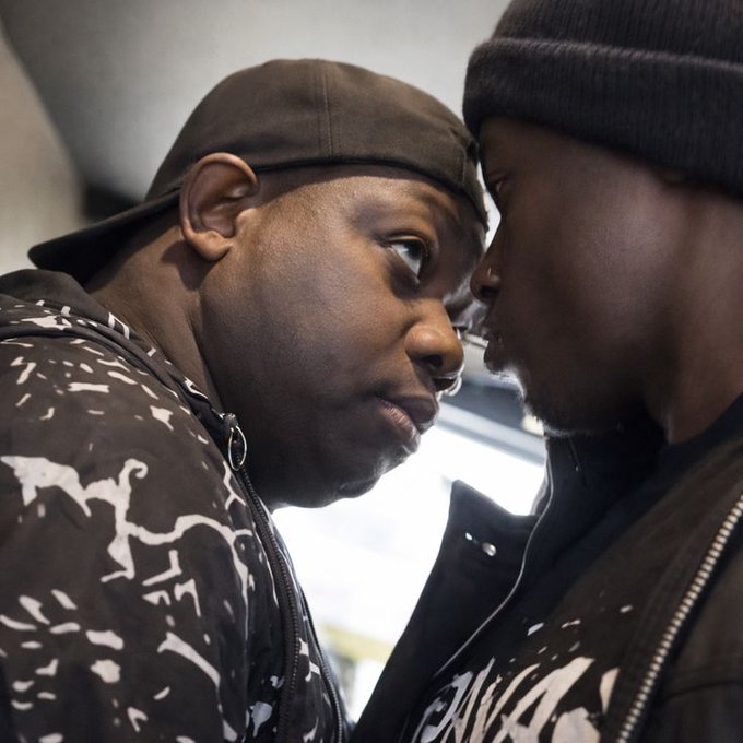 Two men in dark clothing standing face-to-face in close proximity, one wearing a cap and a black-and-white patterned hoodie.