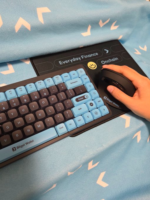 A blue and brown Bitget Wallet keyboard paired with a Bitget mouse rests on a matching branded mat showing labels like "Everyday Finance," "Onchain," and "Bitget Wallet." The coordinated setup screams Bitget Wallet fandom without a single word, perfectly matching the post's challenge.