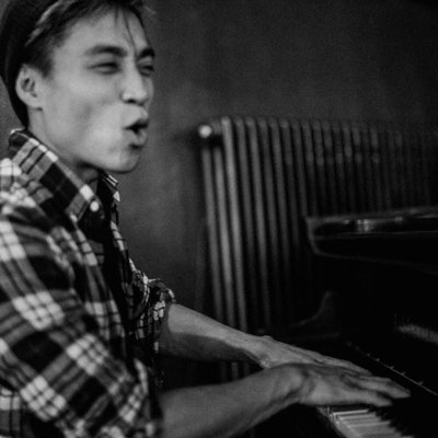 A black-and-white photo of a young man wearing a plaid shirt, winking and pursing his lips while playing a piano next to a radiator.