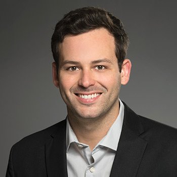 A professional headshot of a smiling young man with short dark hair, wearing a black suit jacket and light gray dress shirt against a dark gray studio background.