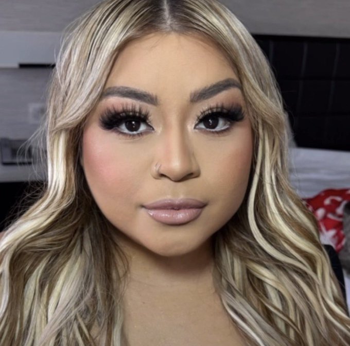Close-up selfie of a woman with long wavy blonde hair, heavy eye makeup, long lashes, and glossy pink lipstick.