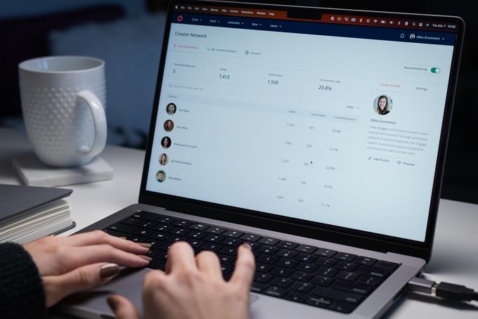 A person's hands type on a laptop keyboard displaying an Etsy "Creator Network" dashboard showing 7,413 visits, 1,545 subscribers, and a 22.8% conversion rate, with a white mug beside the laptop.
