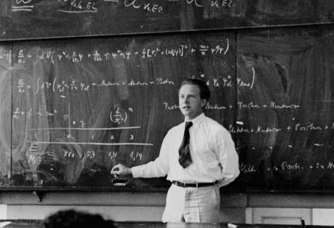 A black and white photograph of a man in a white shirt and tie standing in front of a chalkboard filled with physics equations and calculations, appearing to be a scientist or professor giving a lecture.