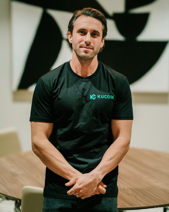 The image features a man wearing a black t-shirt with the word KuCoin written across his chest. He has his hands crossed and is standing next to a dining table. The background of the image shows a painting, which adds an artistic touch to the scene. The caption for this image reads "We love to see our Managing Director of KuCoin in @kucoinaustralia James Pinch, James Pinch, taking the stage at #DECon2026!".