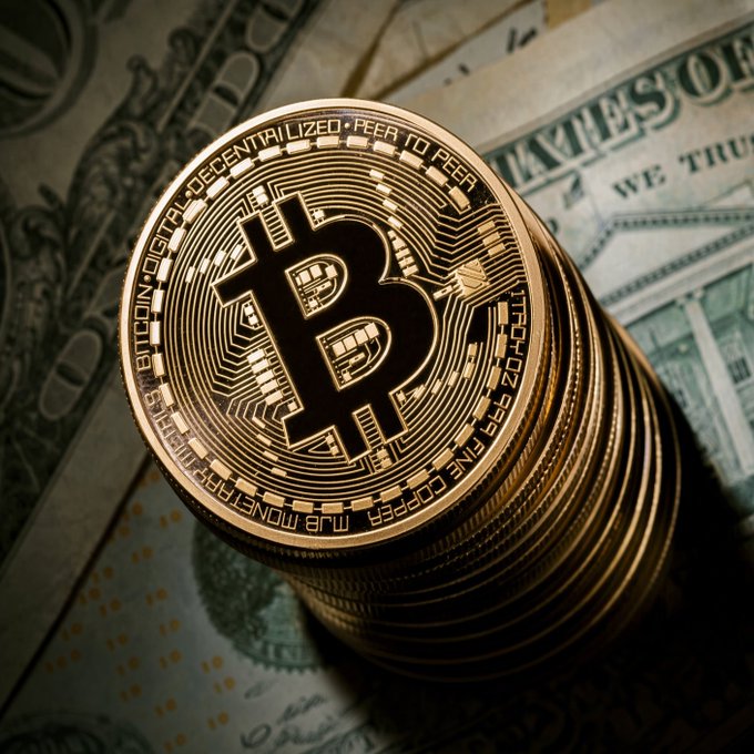 A stack of gold-colored Bitcoin coins resting on US dollar bills, with the top coin showing the Bitcoin 'B' logo and inscriptions including 'BITCOIN DIGITAL DECENTRALIZED PEER TO PEER' and 'FINE COPPER MJB MONETARY METALS'.