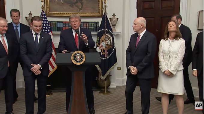 President Donald Trump stands at a podium bearing the presidential seal in the White House, flanked by officials including David Sacks and several business executives, as he addresses attendees of his crypto summit meeting. The image illustrates the "political show" described in the post, capturing the formal White House setting where Trump promoted his administration's support for the cryptocurrency industry ahead of the midterm elections.
