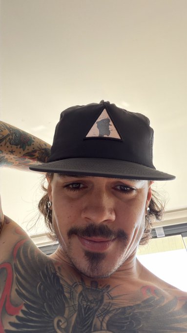 A man wearing a black snapback cap with a Bored Ape Yacht Club (BAYC) emblem on the front is shown, with visible tattoos on his arm and chest. The post references "M1 Bayc flipped brim," referring to the M1 BAYC style snapback with a flat/folded brim, aligning with the cap in the image.