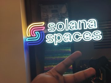 Photo of a person making a peace sign in front of a neon sign that reads 'solana spaces' with the colorful Solana logo.