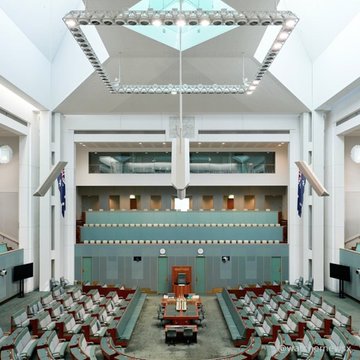 An interior view of the Australian House of Representatives chamber, featuring green seating arrangements arranged in rows facing a central speaker's chair beneath a modern lighting array.