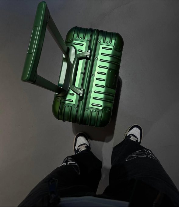 A green hard-shell suitcase with an extended telescoping handle stands on a gray floor, viewed from above as someone in black pants and white sneakers stands next to it.