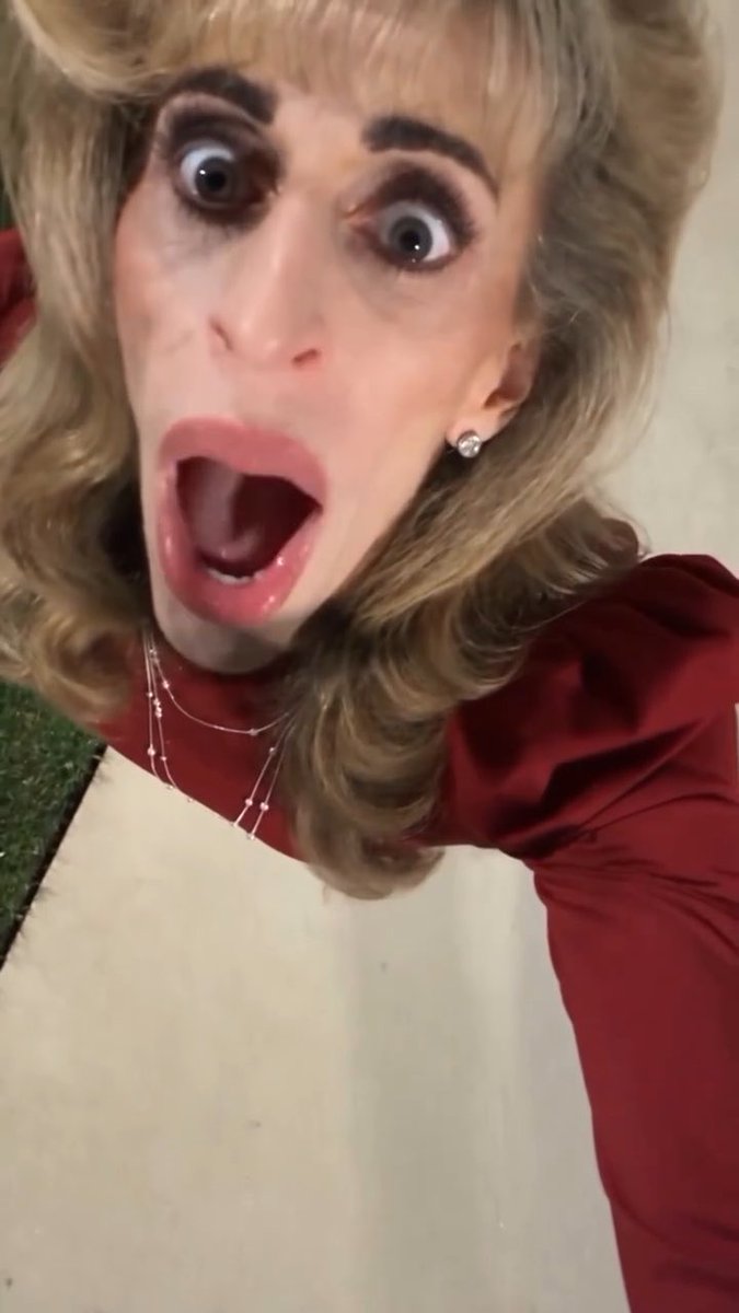 **Dramatic Makeup Look:** A close-up, high-angle view of a person with a shocked expression, wide eyes, and open mouth, wearing heavy makeup, a blonde wig, and a red top.