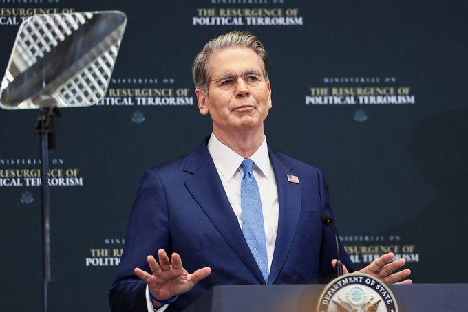 A man in a blue suit and tie stands at a podium with the Department of State seal, with the backdrop reading 'MINISTERIAL ON THE RESURGENCE OF POLITICAL TERRORISM'.