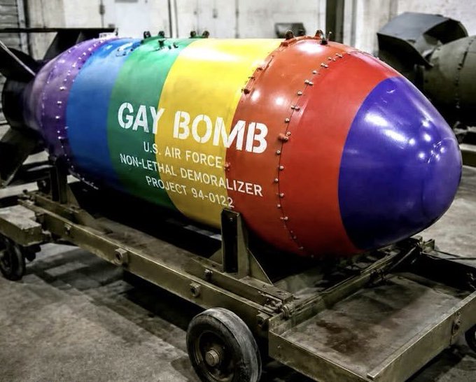 A rainbow-colored bomb labeled 'GAY BOMB - U.S. AIR FORCE - NON-LETHAL DEMORALIZER - PROJECT 9A-0122' sits on a metal cart in what appears to be a warehouse.