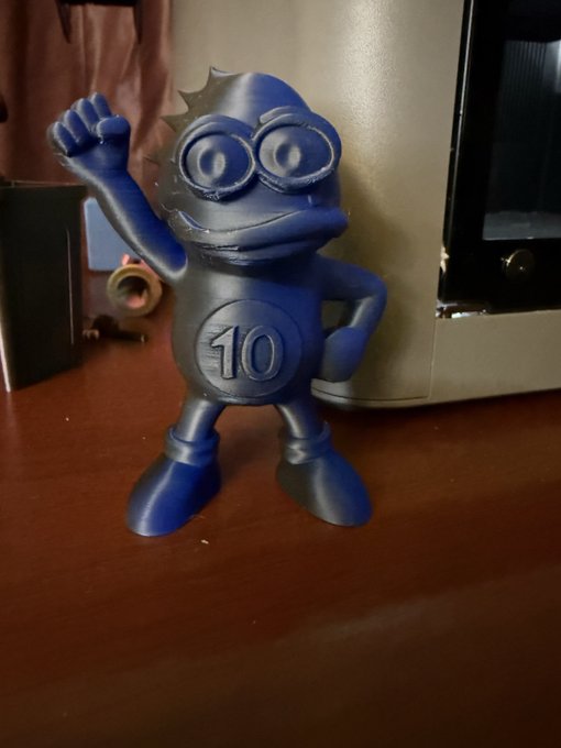 A blue plastic figurine with "10" emblazoned on its chest stands on a wooden desk, raising one hand in a cheerful wave—appearing to be a character from a casual gaming context. The caption "a wild @ItsCrapto appears" humorously frames the toy as if it just spawned in the wild, mimicking Pokémon-style encounters.