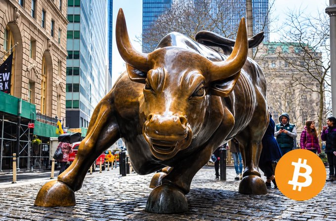 The bronze Charging Bull statue in New York City's Financial District with a Bitcoin logo overlaid in the bottom right corner.