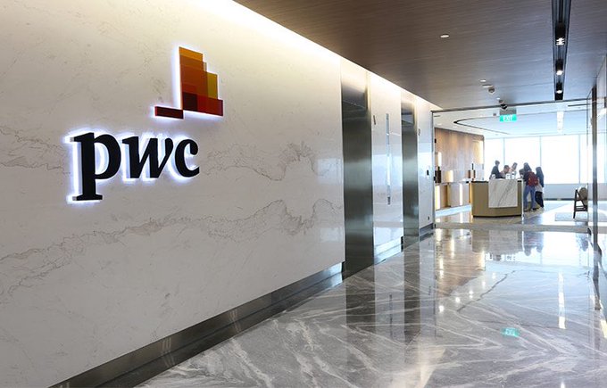 A modern office lobby hallway with a marble floor, featuring a lit "pwc" logo on a white marble wall and an elevator down the hall.