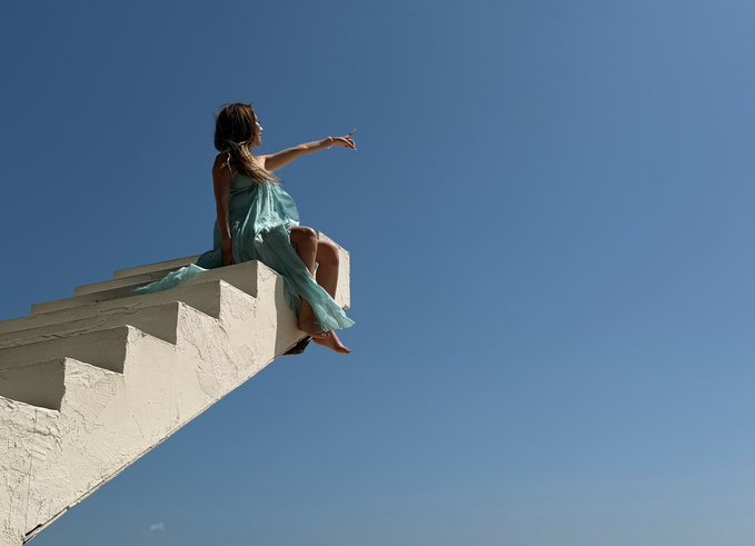 A woman in a flowing teal dress sits perched on the edge of a steep concrete staircase, pointing into the vast blue sky below her feet.

The image perfectly captures the precarious, high-stakes feeling of the post's lament about crypto trading—balancing on the knife's edge with profits and losses hanging in the balance, much like sitting at the tip of a staircase with nothing but thin air beneath.