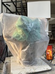 A green, sculpted figure (resembling Yoda) wrapped in clear plastic bubble wrap, sitting on a base in a cluttered workspace.