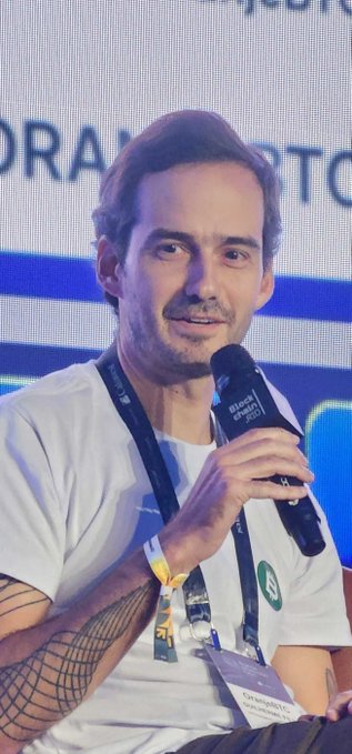 A man with dark hair and a mustache speaks into a microphone at a conference, wearing a white t-shirt and a lanyard, with a 'STAND WITH CRYPTO' lanyard and event branding visible.