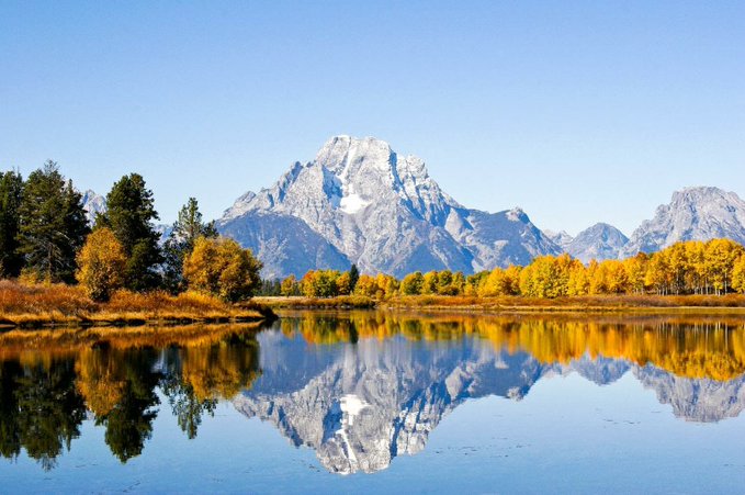 Landscape photograph of snow-capped mountains reflected in a calm lake, bordered by autumn trees with golden foliage under a clear blue sky.