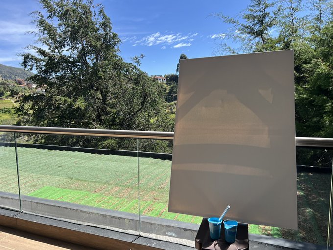 A blank canvas sits on an easel overlooking a lush green field and tree-lined hillside from a balcony in Medellín, with small blue paint cups ready at its base. The setup captures the beginning moments of the artist's residency work, with the vibrant landscape ahead hinting at the colorful Silletera-inspired painting to come.