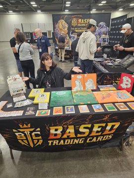 A vendor sits behind a "BASED TRADING CARDS" booth at the Midwest Bitcoin & Blockchain Conference (MWBTCSummit) in Columbus, Ohio, displaying colorful trading cards and merchandise during the second day of the art gallery. The BTC logo and "Based Trading Cards" branding are prominently featured on the table banner.