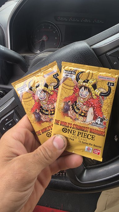A hand holds two OP-17 One Piece booster packs from The World's Strongest Warriors series (OP-17) featuring Bandai Namco branding, photographed inside a car with the steering wheel and dashboard visible in the background.