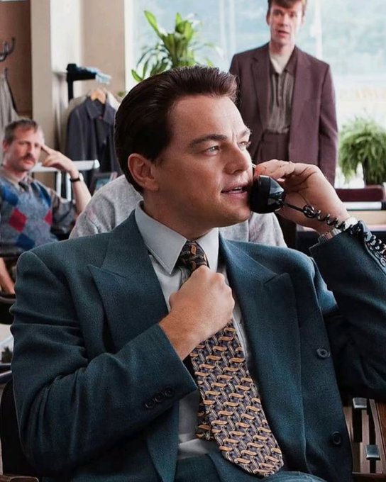 Leonardo DiCaprio sits in a suit while talking on an old corded phone, adjusting his tie, in what appears to be a scene from The Wolf of Wall Street.