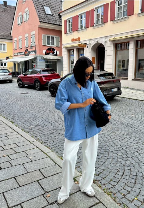 A woman wearing an oversized blue shirt and white pants stands on a cobblestone street in front of European-style buildings.