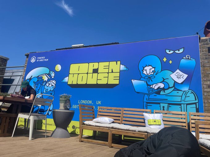 An outdoor deck area with wooden seating and a large blue banner reading 'OPEN HOUSE LONDON, UK' featuring cartoon characters, powered by Arbitrum.