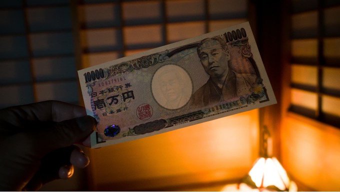 A hand holds up a Japanese 10,000 yen note featuring the portrait of Fukuzawa Yukichi, illuminated against a warm wooden background. The image ties to Japan's bold push to digitize its massive bond market on the blockchain, contrasting the country's traditional paper currency with its forward-looking financial innovation.