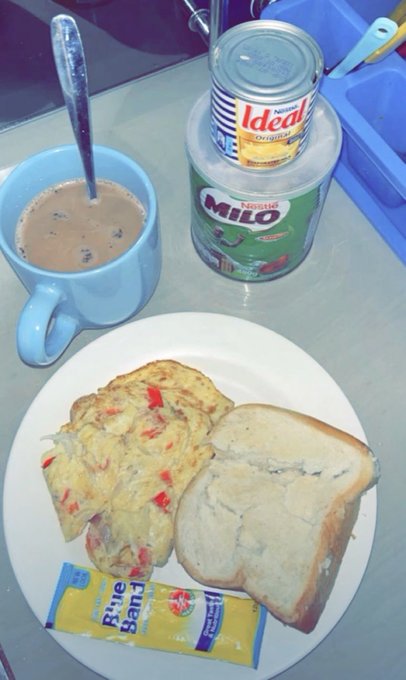 A breakfast spread on a plate includes an omelet with vegetables, a slice of bread, and a 'Bilie Band' candy, accompanied by a cup of coffee, a tin of Ideal condensed milk, and a can of Nestle Milo.