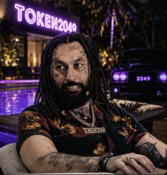 A man with dreadlocks, facial tattoos including '2049', and grillz, wearing a floral shirt, seated near a neon 'TOKEN2049' sign.