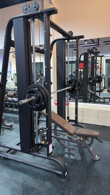 A Smith machine weight rack equipped with a bench, barbell, and weight plates stands in a gym with mirrors on the wall.